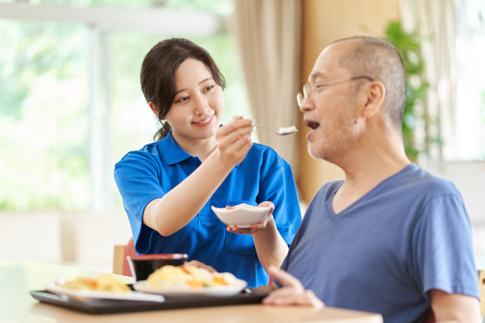 Caregiver feeding elderly man in dining area.